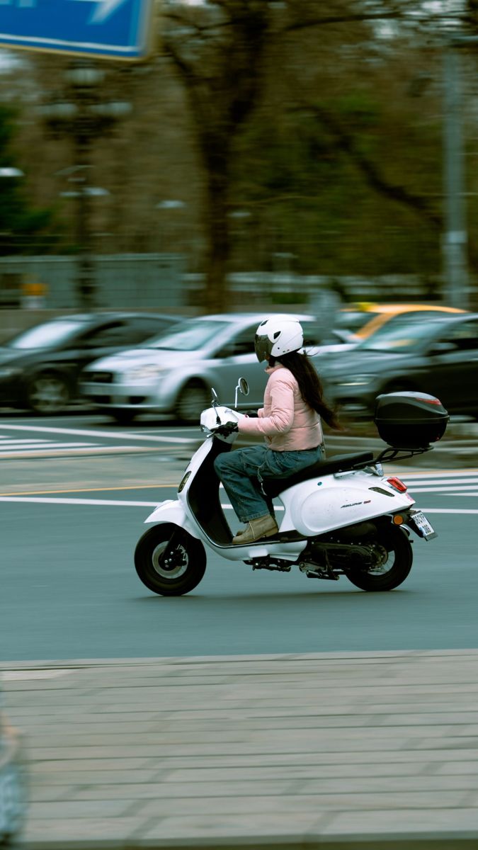 Person in a white helmet riding a white scooter along a road, cars blurred behind